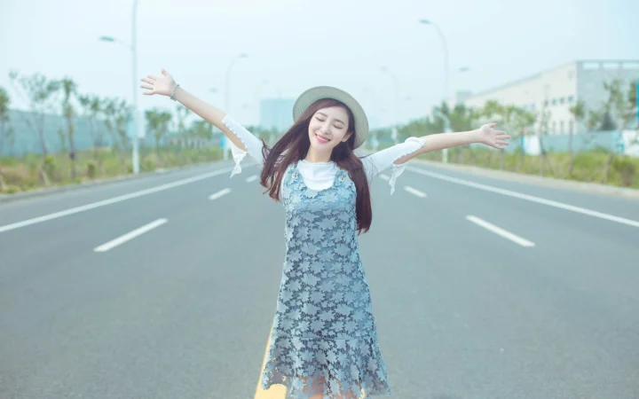一名戴帽子的年轻女子站在空旷公路中央，张开双臂面向镜头微笑。