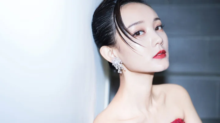 一位盘发并佩戴华丽耳饰的女性以侧脸回望镜头，红唇妆容映衬着浅色背景。
