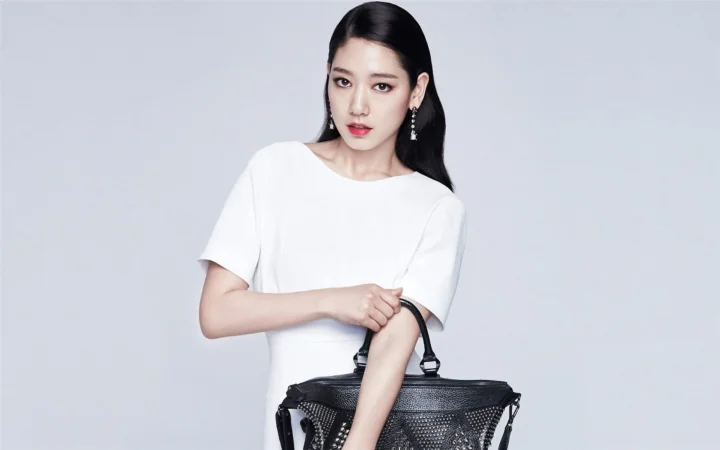 흰색 상의를 입은 여성이 연한 회색 배경 앞에서 검은색 핸드백을 들고 있다.