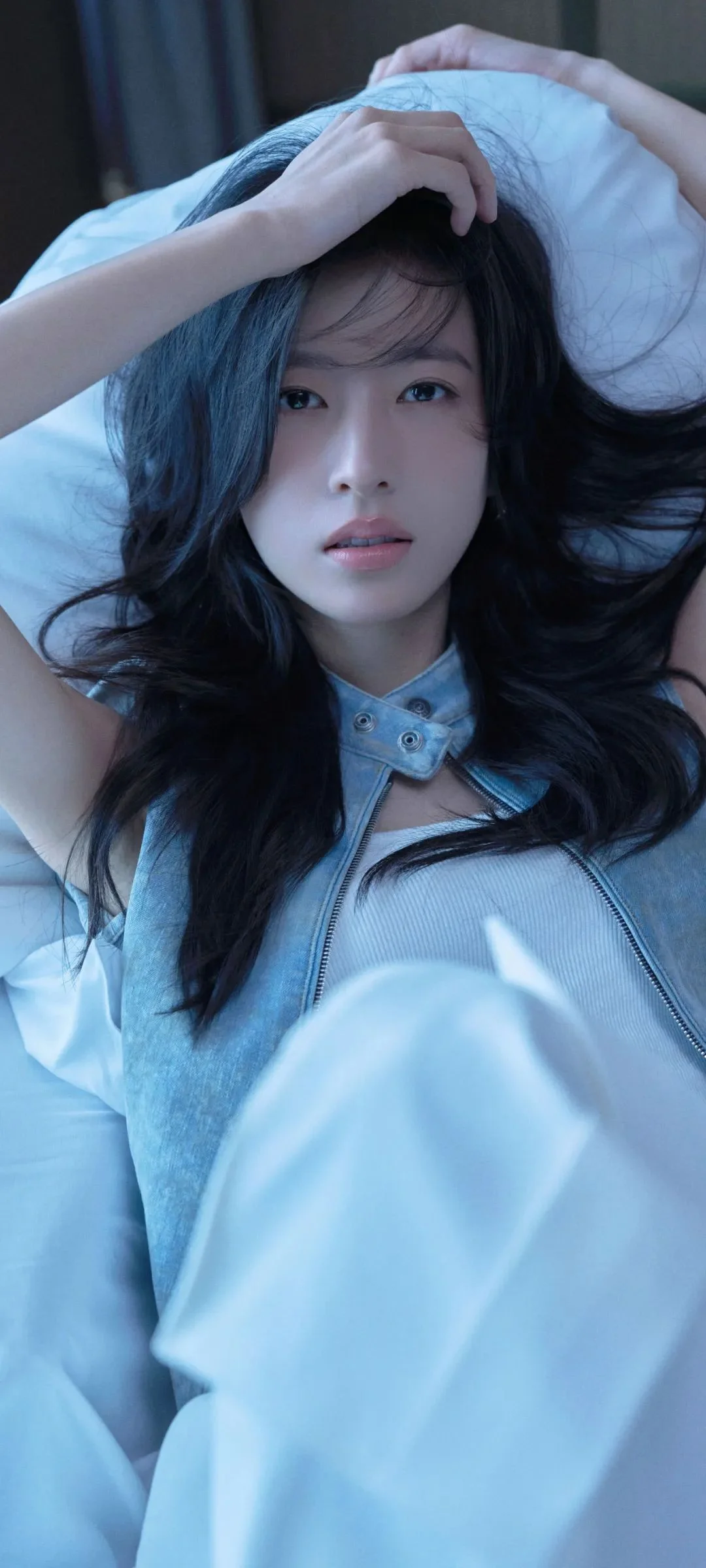 長い髪の女性が明るい色のベッドに横たわる、青みがかったポートレートです。