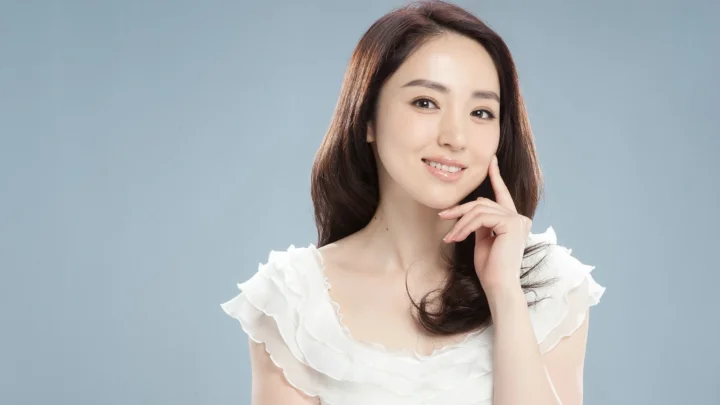 흰색 러플 상의를 입은 성인 여성이 연한 청회색 배경 앞에서 손을 뺨에 대고 미소 짓고 있습니다.