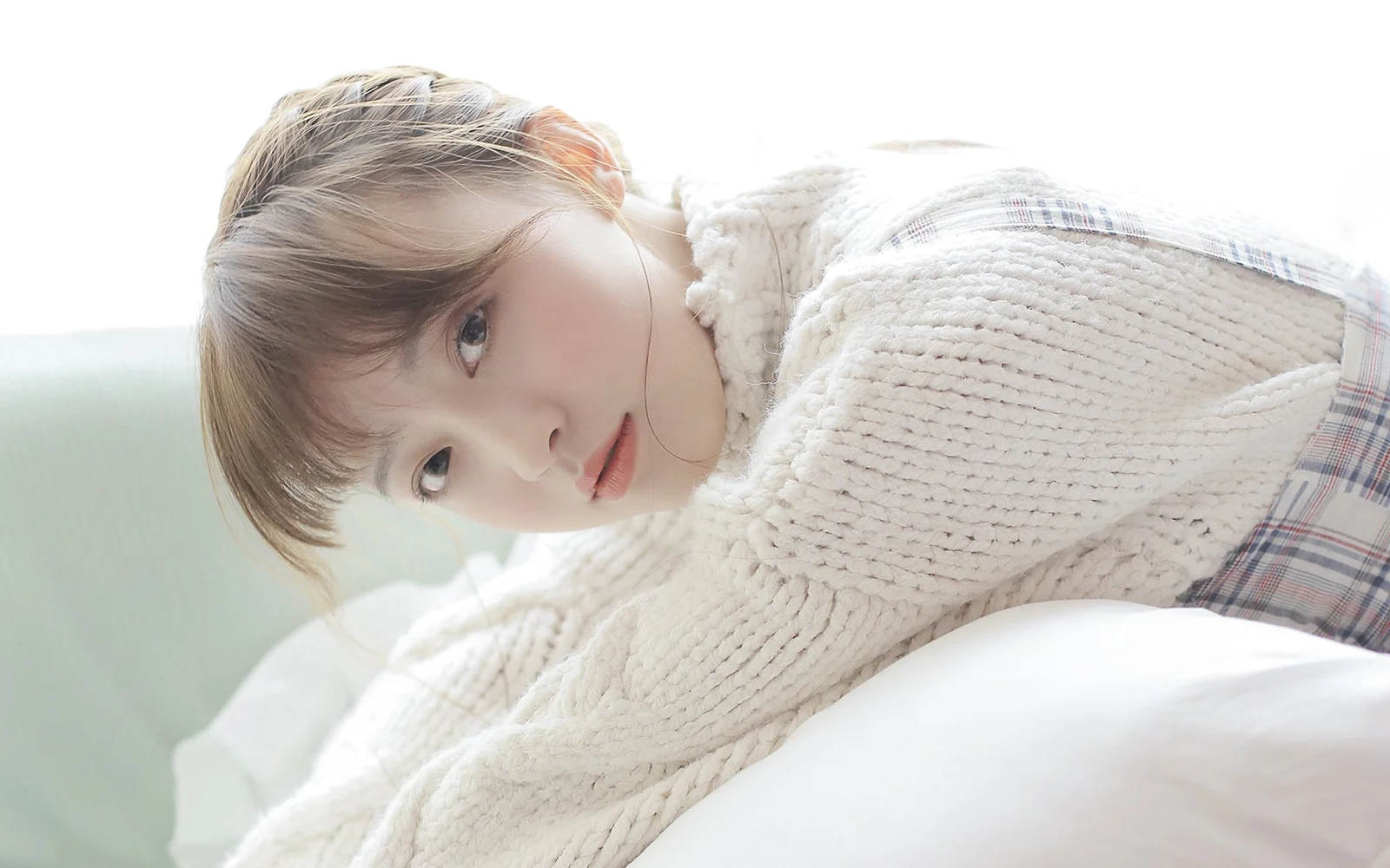 밝은 색 침대에 누운 아이보리색 니트 스웨터 차림의 성인 여성이 카메라를 바라보고 있습니다.
