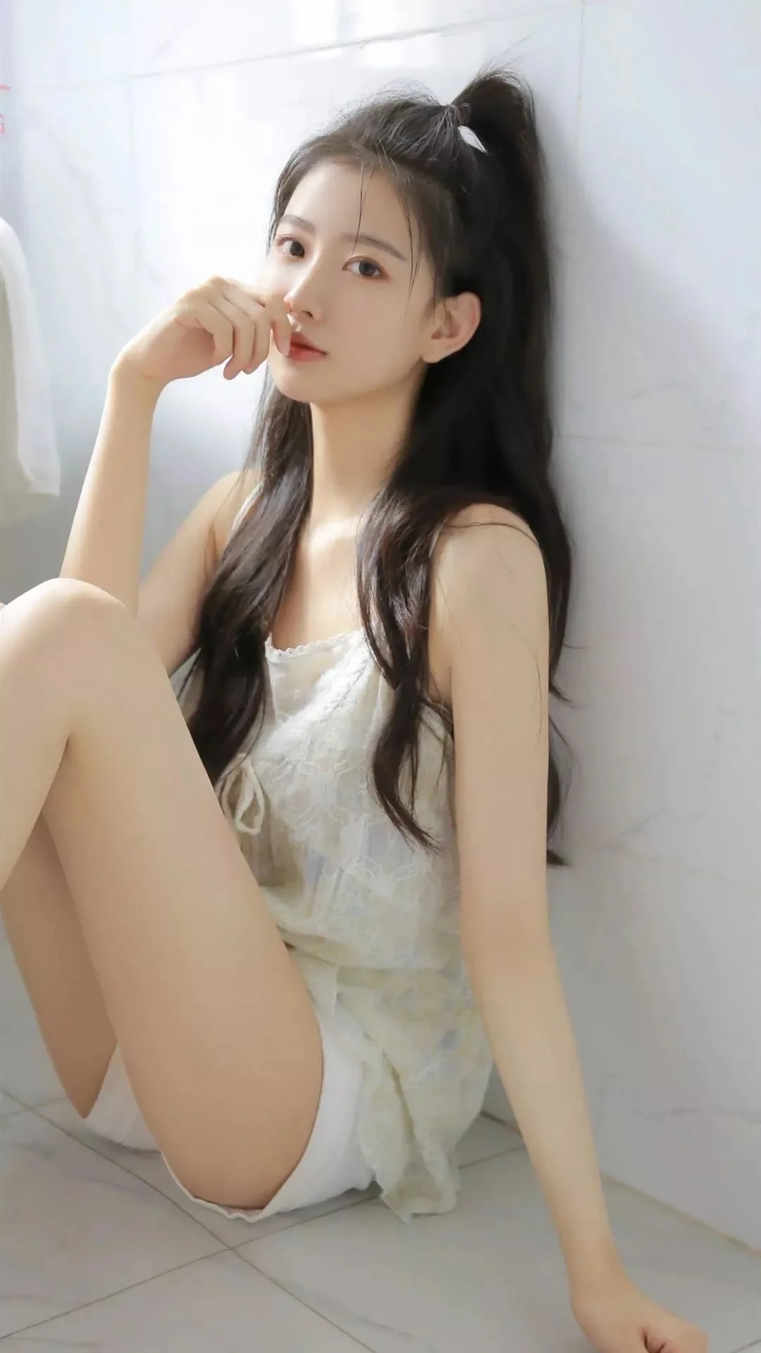 一名長髮女子坐在室內白色牆壁旁，穿著白色服裝並看向鏡頭。