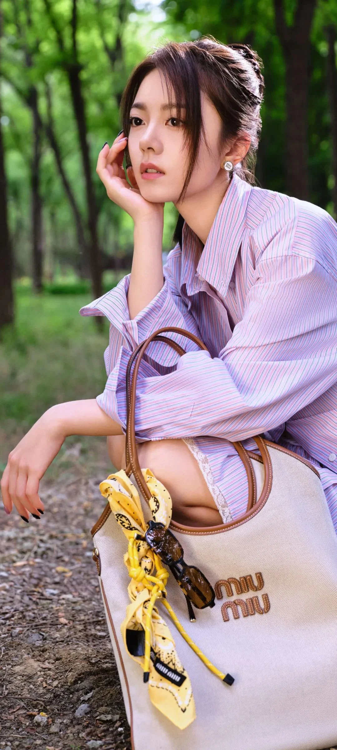 女性が森の小道のそばに座り、頬に手を添え、隣に淡い色のハンドバッグを置いている。