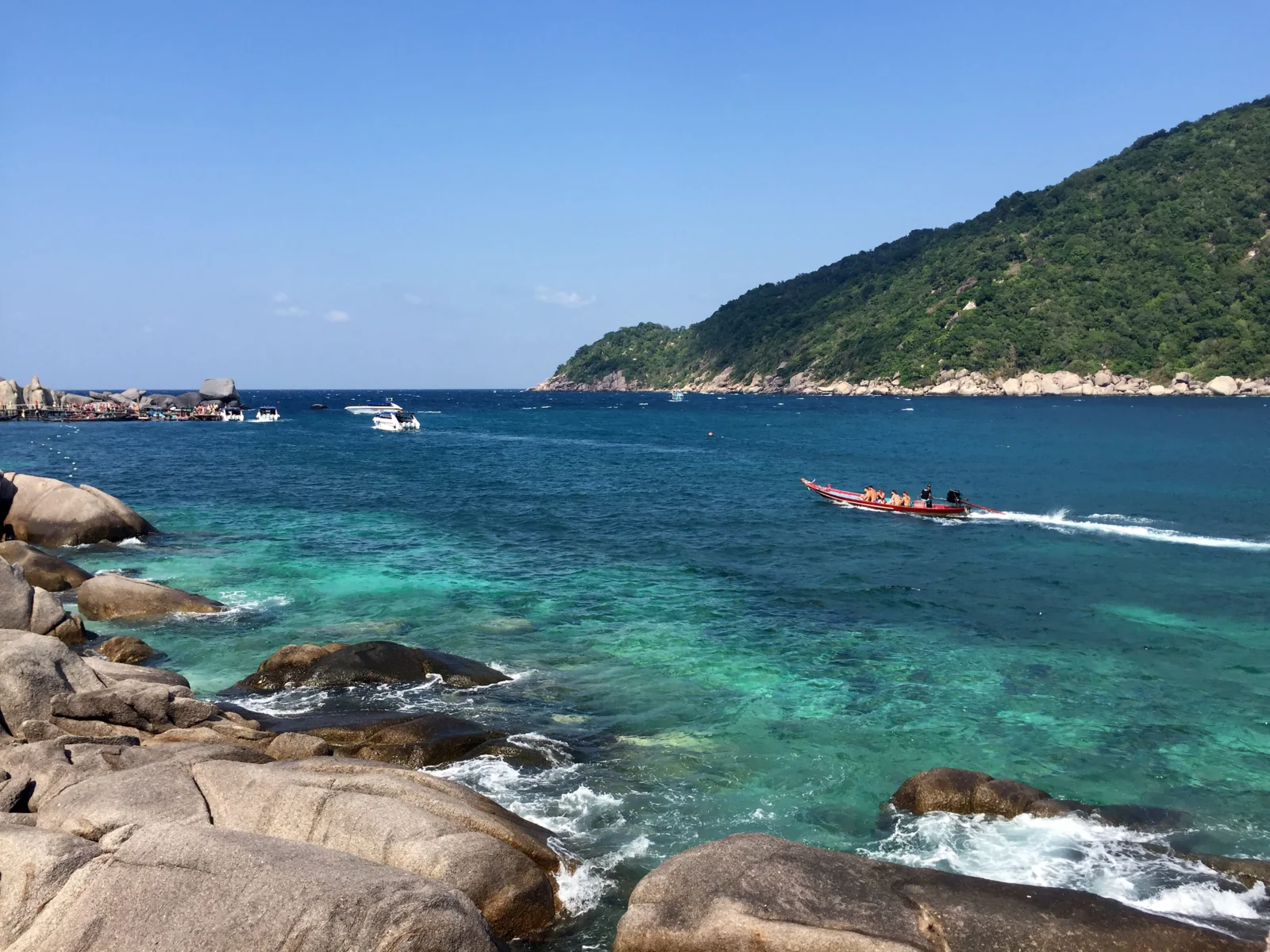 푸른 하늘 아래 투명한 바다와 바위 해안, 초록빛 언덕, 한 척의 보트가 어우러진 평온한 해안 풍경입니다.