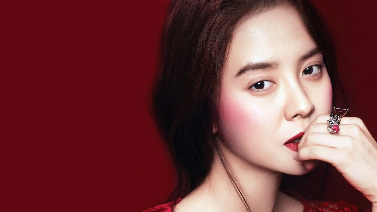 짙은 빨간색 배경 앞에서 성인 여성이 손가락을 입술 가까이에 대고 카메라를 바라보고 있습니다.