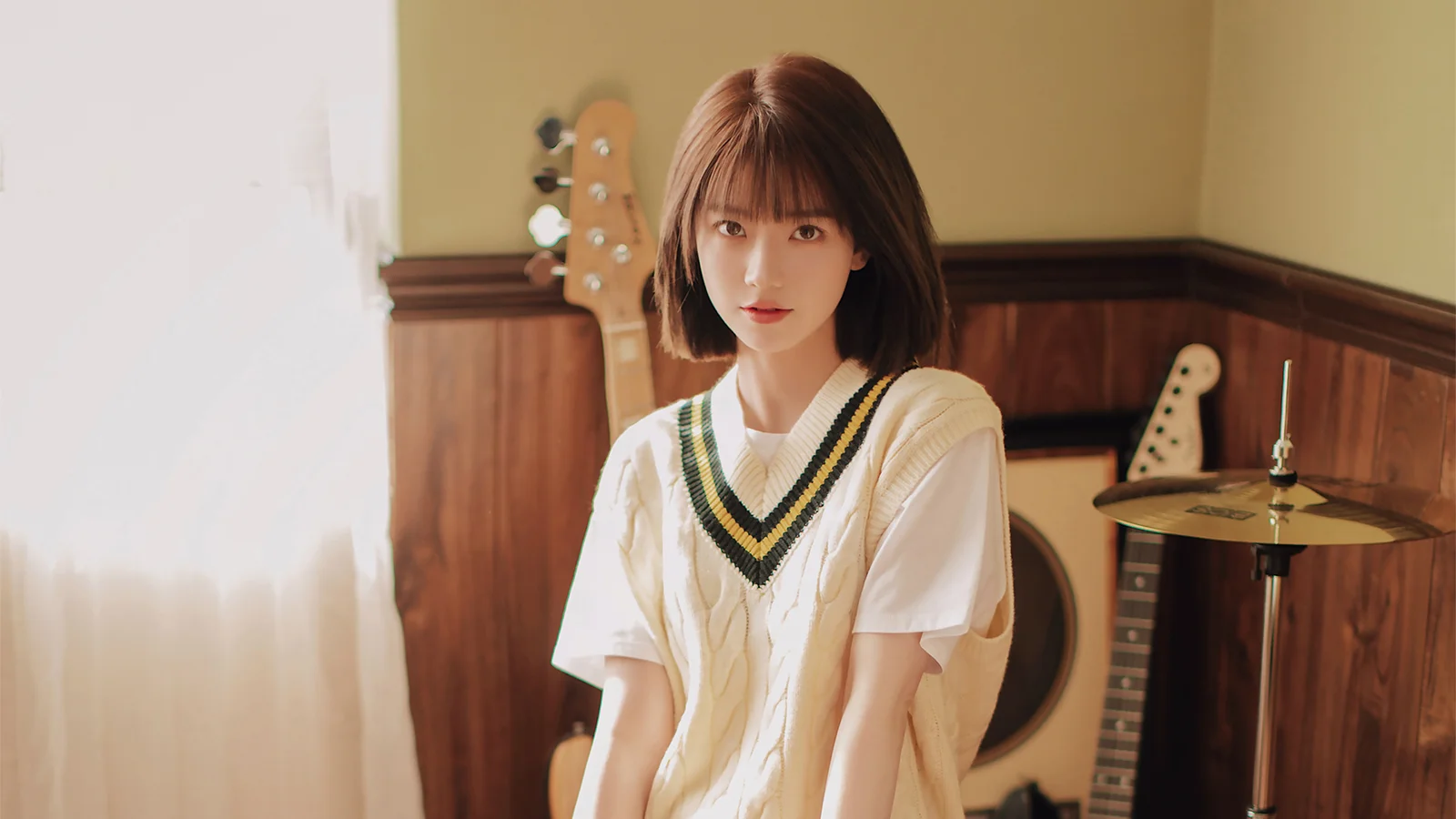 나무 벽으로 꾸며진 실내 음악 공간에서 기타와 드럼 세트를 배경으로 여성이 앉아 있습니다.