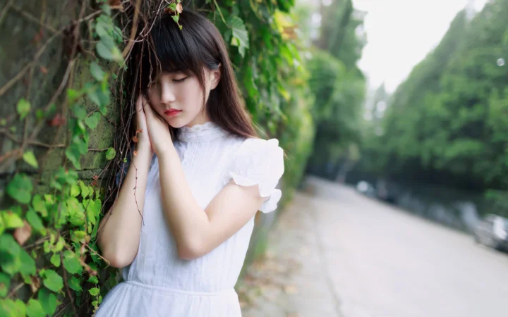 白いワンピースを着た女性が、蔓植物のそばに立ち、背景には木々に囲まれた道が見える。