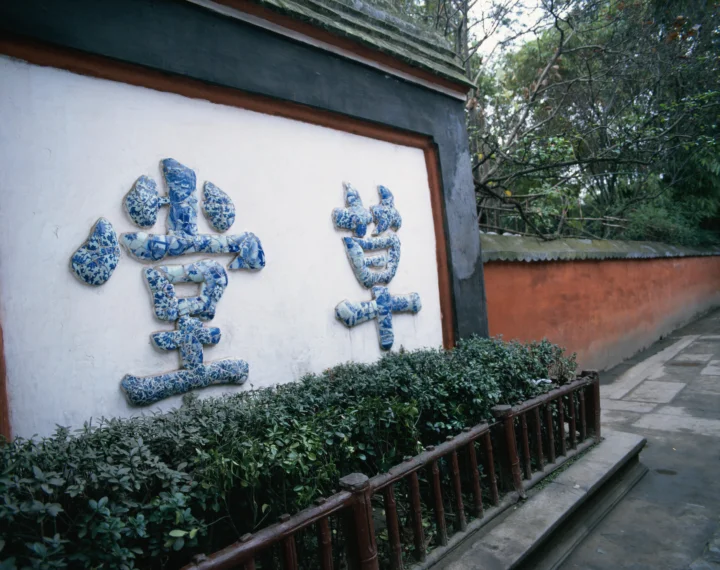 白色古墙上镶嵌着蓝白瓷片书法，前方是绿篱、木栏和石板小径。