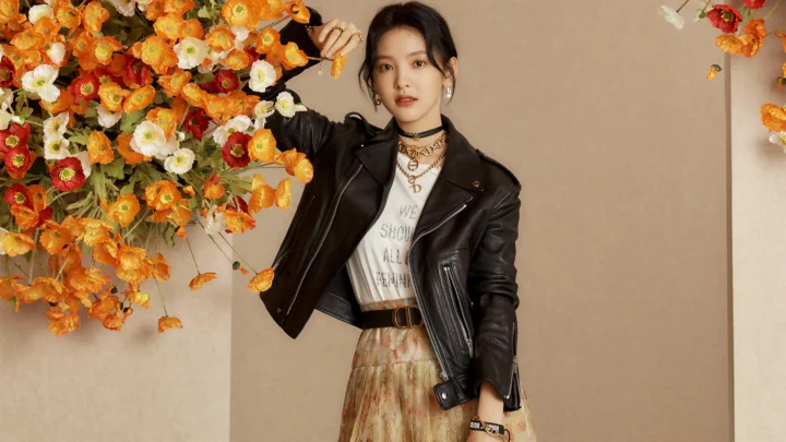 검은색 가죽 재킷을 입은 성인 여성이 주황색과 흰색 꽃을 배경으로 서 있다.