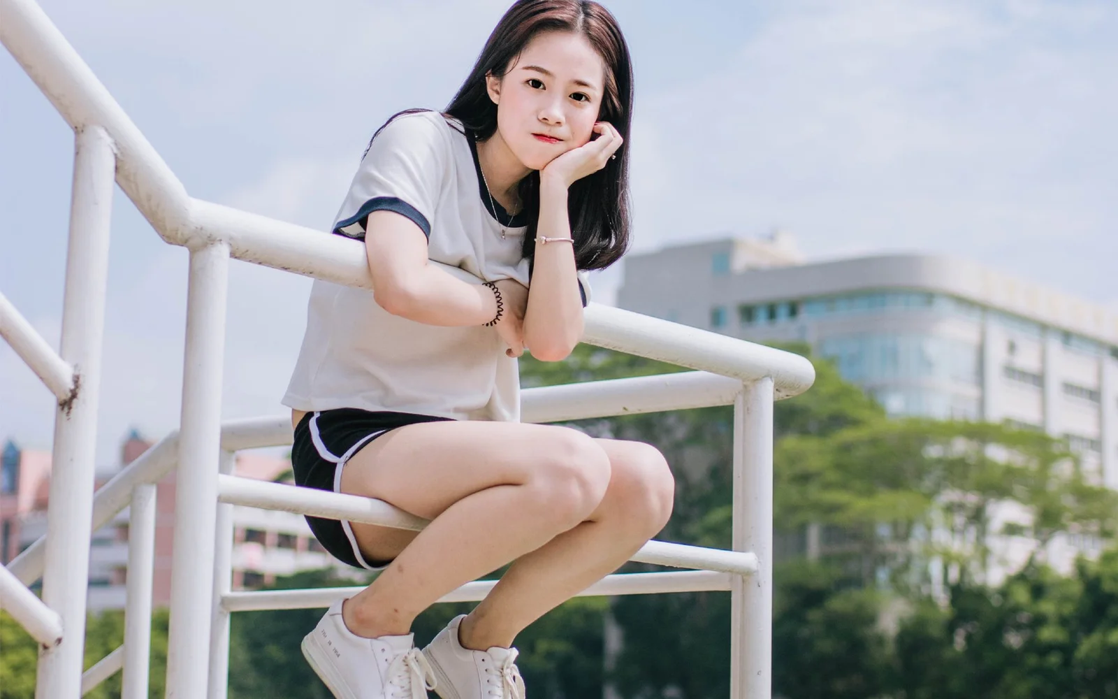 一名年輕女性坐在戶外白色欄杆上，背景是樹木和城市建築。