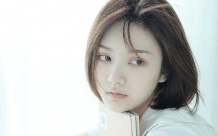 밝은 배경 앞에서 갈색 단발머리의 성인 여성이 밝은 색 책을 들고 있는 클로즈업 인물 사진입니다.