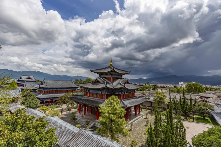 一座中国传统建筑群坐落在群山之间，天空中布满层叠的乌云与白云。