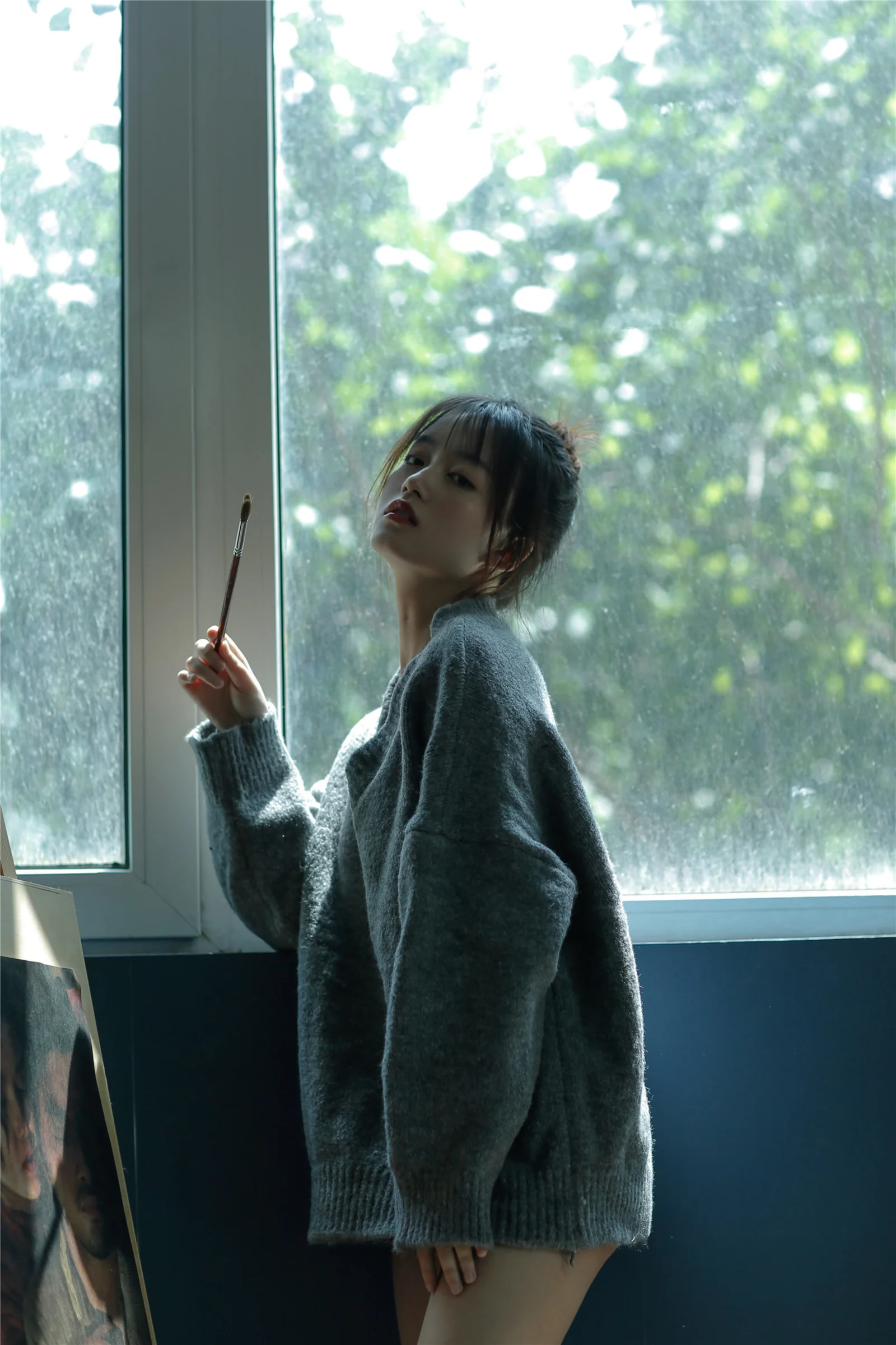 一名身穿灰色毛衣的女性站在大窗户旁，手里拿着一支细画笔。