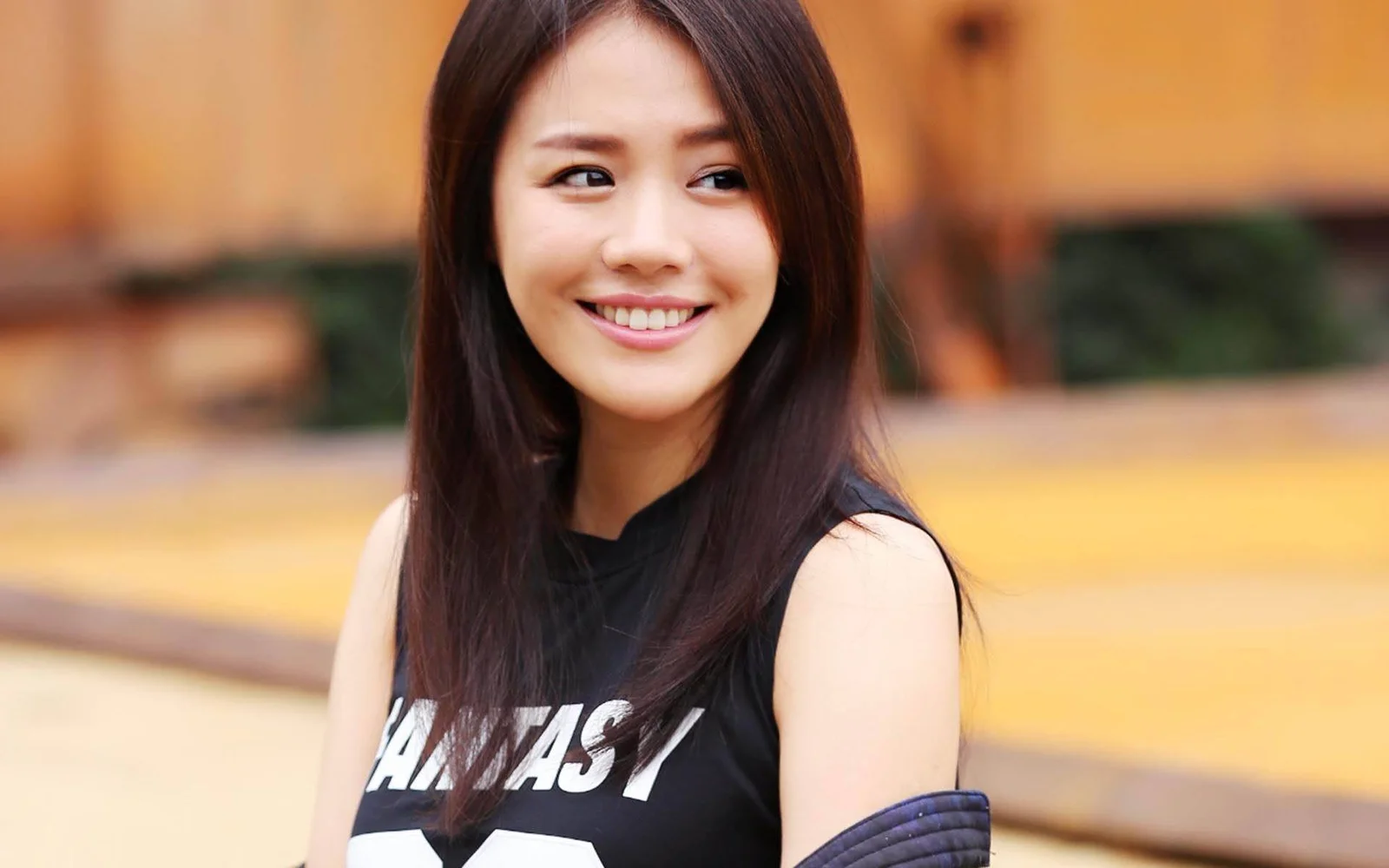 一名长发女性穿着黑色无袖上衣，在户外背景前微笑看向一侧。