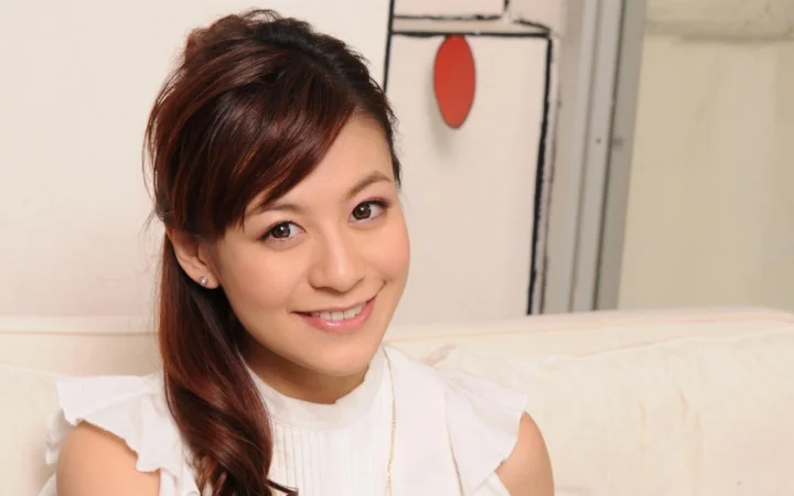 一位棕色长发女性穿着白色上衣，坐在浅色沙发上微笑面对镜头。