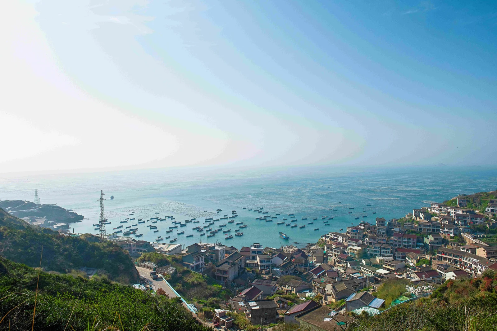 緑豊かな斜面から、漁船が浮かぶ青い湾と海岸沿いの集落を見下ろしている。