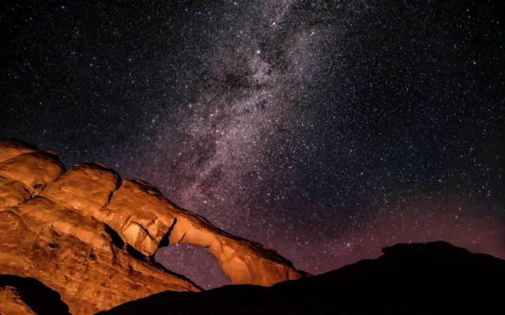 璀璨的银河横贯夜空，照亮前景中的橙色岩石拱门与黑色山脊。