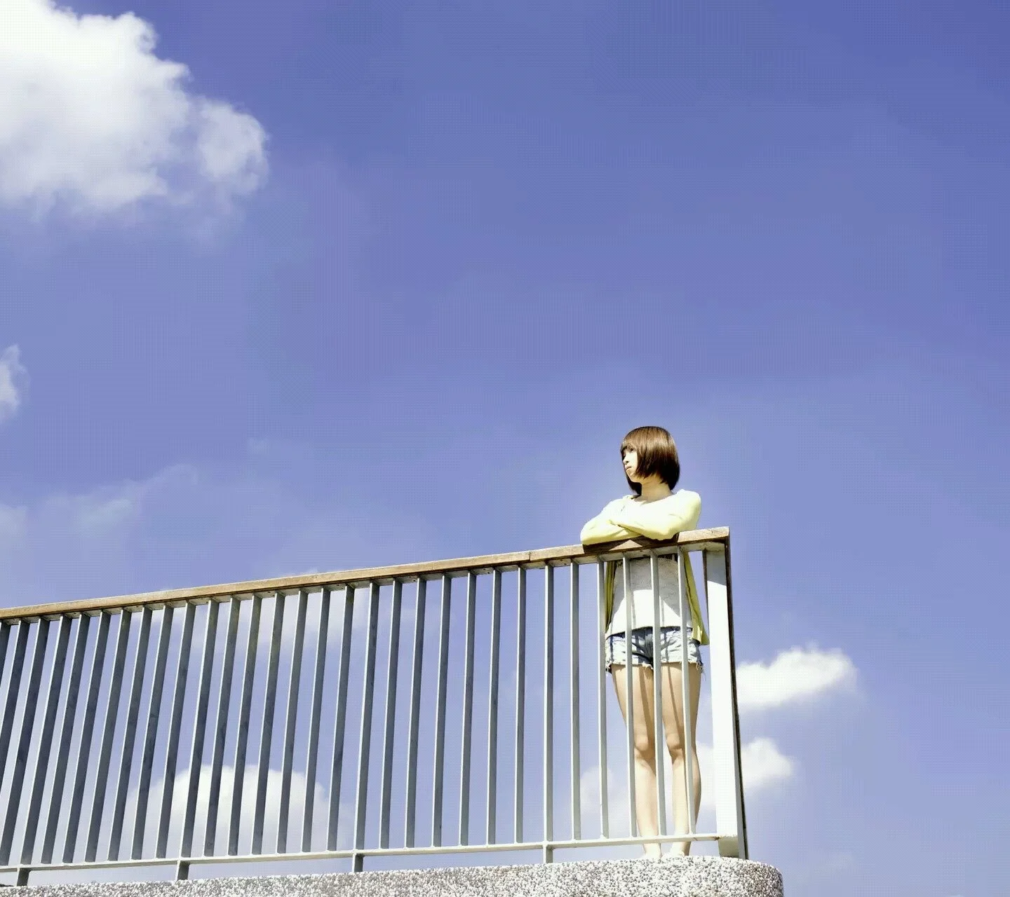 青空の下、女性が白い雲の浮かぶ空を背景に手すりのそばに立っています。