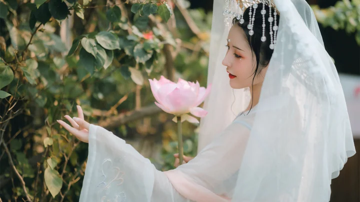흰색 전통 의상을 입은 여성이 푸른 정원에서 분홍색 연꽃을 들고 있습니다.