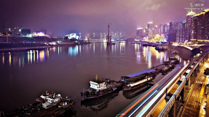 夜色中的城市沿江而立，河面倒映着五彩灯光，前景道路上的车流形成明亮光轨。