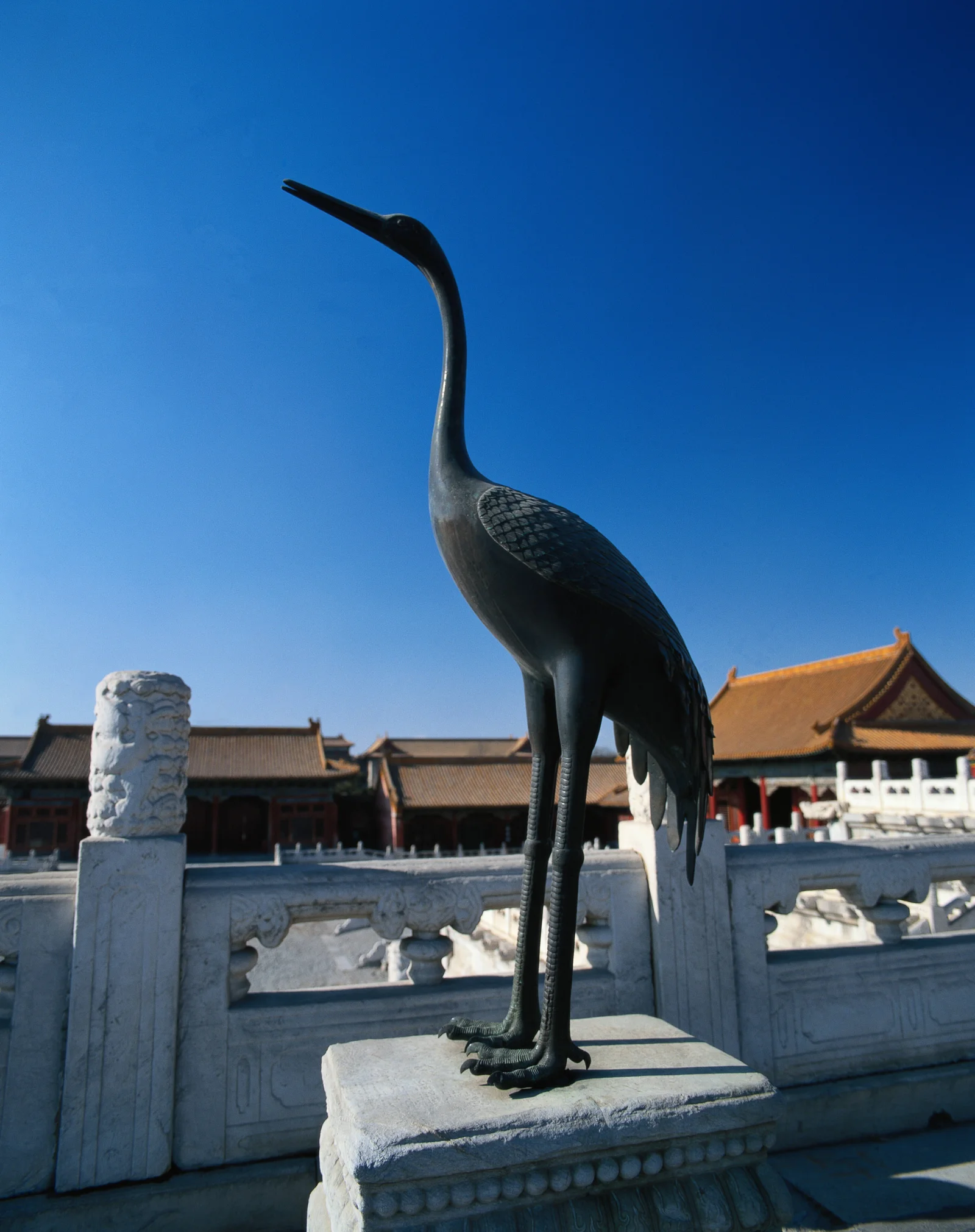 一只青铜鹤雕像矗立在宫殿建筑与湛蓝天空前方。