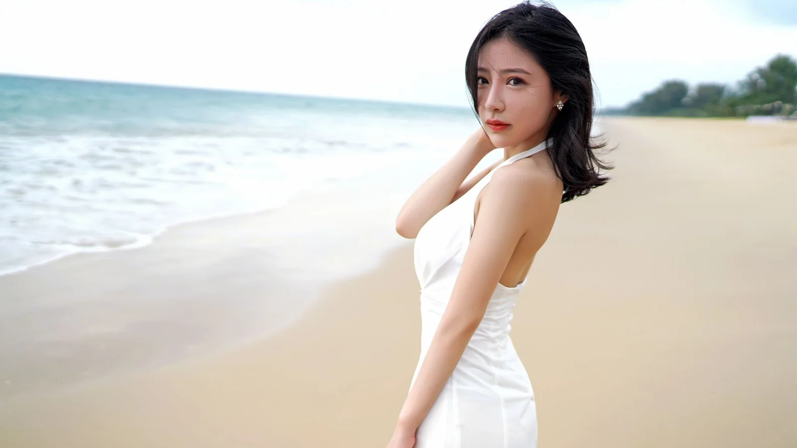 一名穿白色连衣裙的女子站在海滩上，身后是海水与沙滩。