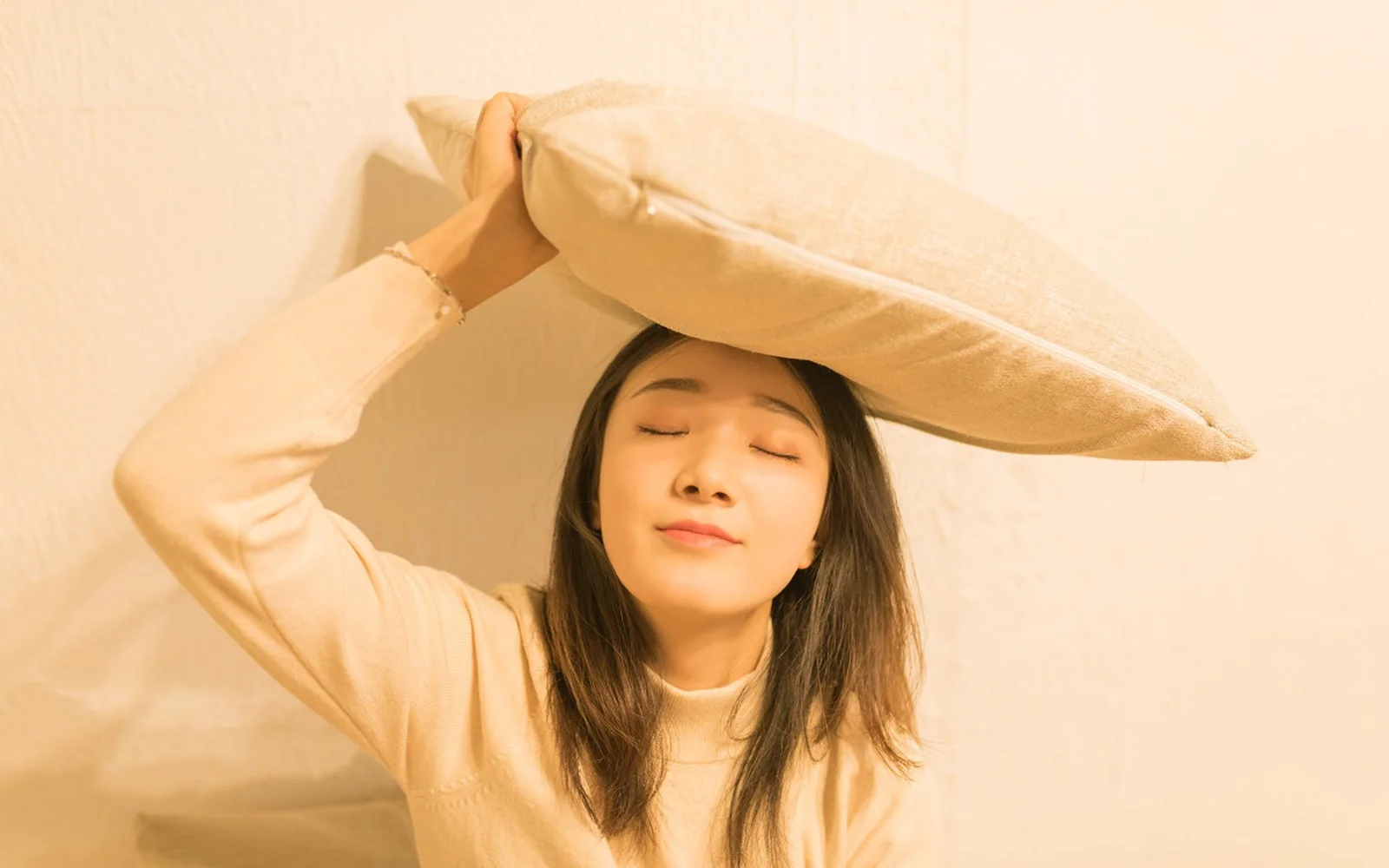 女性が目を閉じ、ベージュの枕を頭上に掲げています。