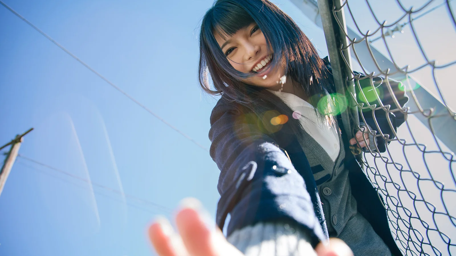一名短发女子在蓝天下靠近铁丝网微笑伸手。