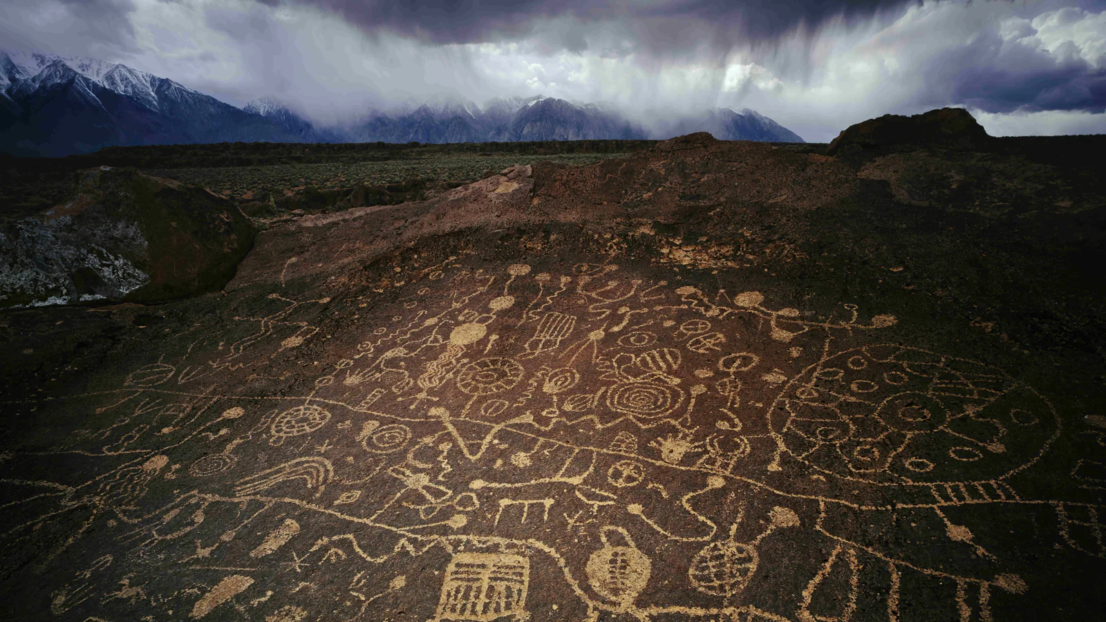 岩石高原上布满古老岩画，远处是群山和厚重的暴雨云层。