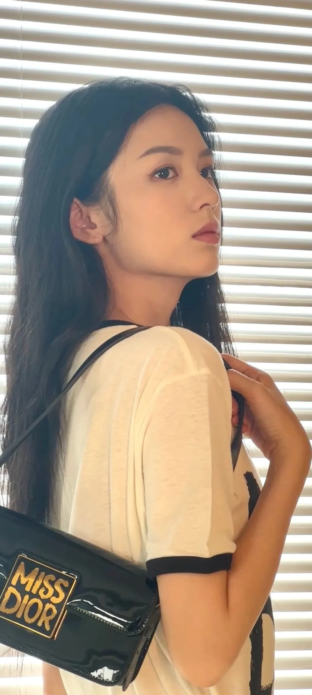 성인 여성이 블라인드 앞에 서서 검은색 가방을 어깨에 메고 카메라 밖을 바라보고 있습니다.