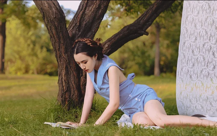 一名身穿浅蓝色连衣裙的成年女子在草地上的树旁俯身。