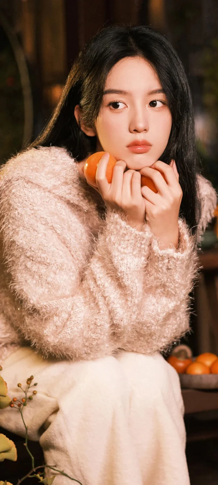 연한 색 스웨터를 입은 여성이 오렌지 옆에서 손으로 턱을 괴고 있습니다.