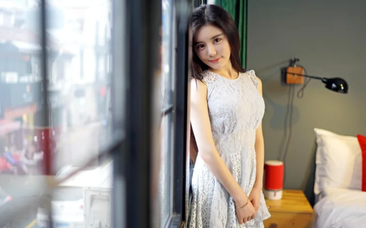 흰색 레이스 원피스를 입은 여성이 침실 창가에 서 있습니다.