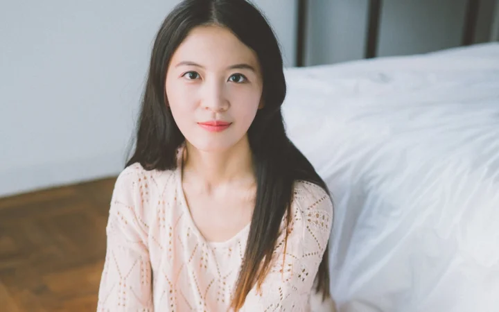 一名长发女子穿着浅粉色针织衫坐在室内床边。