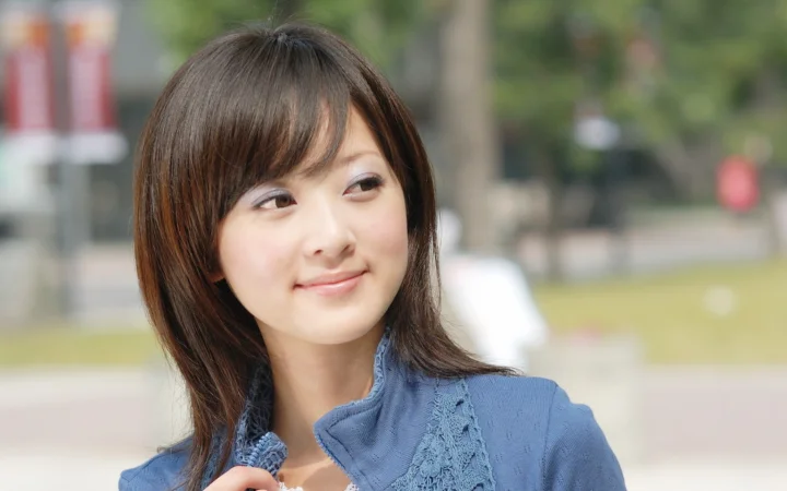 一名棕色长发女子身穿蓝色上衣，在户外背景前微笑侧望。