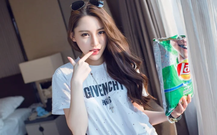 長い髪の女性が白いTシャツを着て窓辺に立ち、お菓子の袋を持っています。