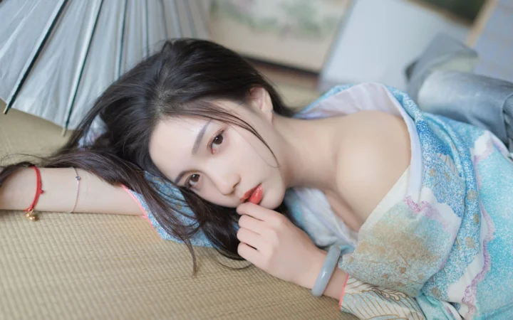 一名长发女性侧躺在编织垫上，手指靠近嘴唇，身旁可见浅色伞面和柔和室内背景。
