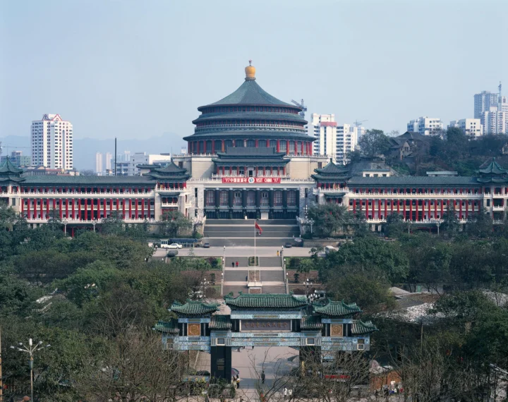 一座大型中式传统建筑坐落在绿树之间，背景是城市高层建筑和晴朗天空。