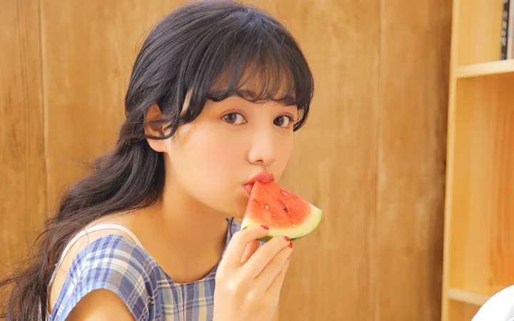 성인 여성이 나무 실내 배경 앞에서 수박 조각을 들고 먹고 있다.