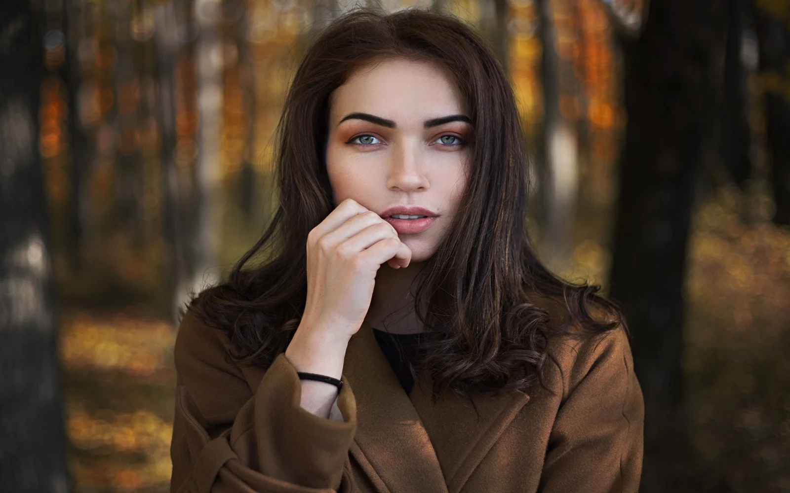 一名長髮女性站在森林中，手靠近嘴唇，背景是柔和的樹木光影。