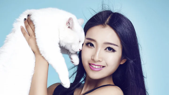 미소 짓는 여성이 흰 고양이를 안고 연한 파란색 배경 앞에 서 있습니다.