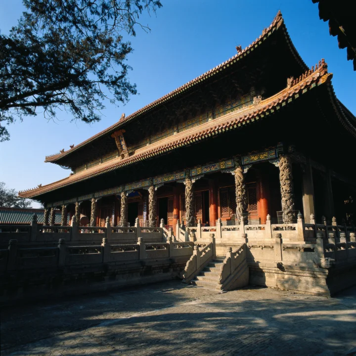 蓝天下，一座带有雕刻石柱和彩色屋檐的中国古建筑坐落在庭院中。