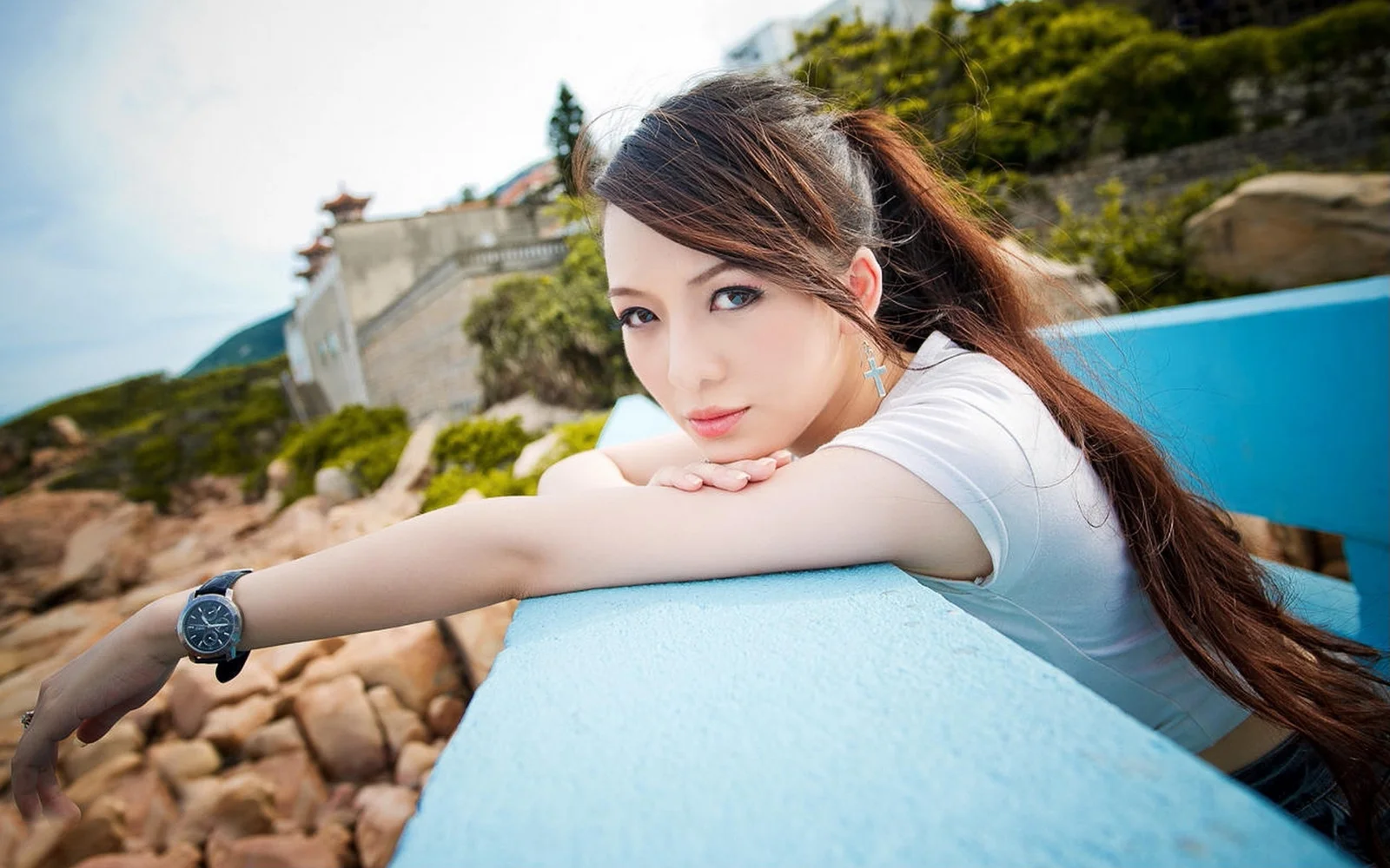 一名女子靠在浅蓝色栏杆上，身后是岩石海岸、树木和建筑。