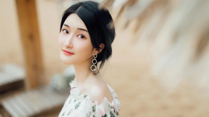 一位黑发女子穿着浅色花卉肩部露肤上衣，在暖色背景前回眸微笑。
