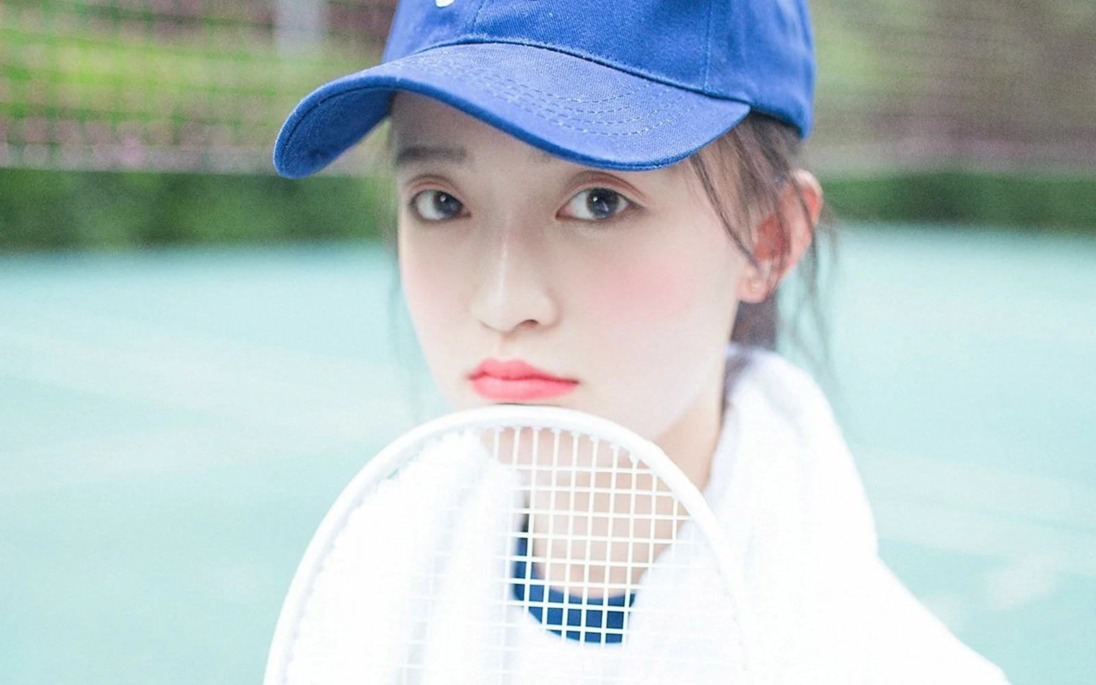 파란 모자를 쓴 여성이 야외 코트 앞에서 테니스 라켓을 들고 있다.