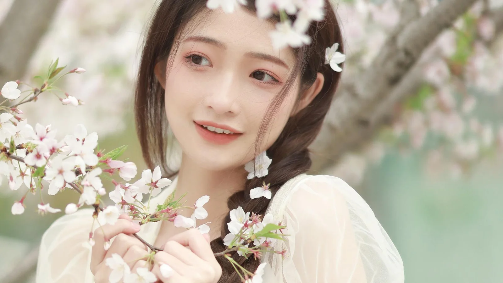 一名女子在盛开的樱花枝旁微笑，背景呈现柔和的浅绿色与粉白色调。