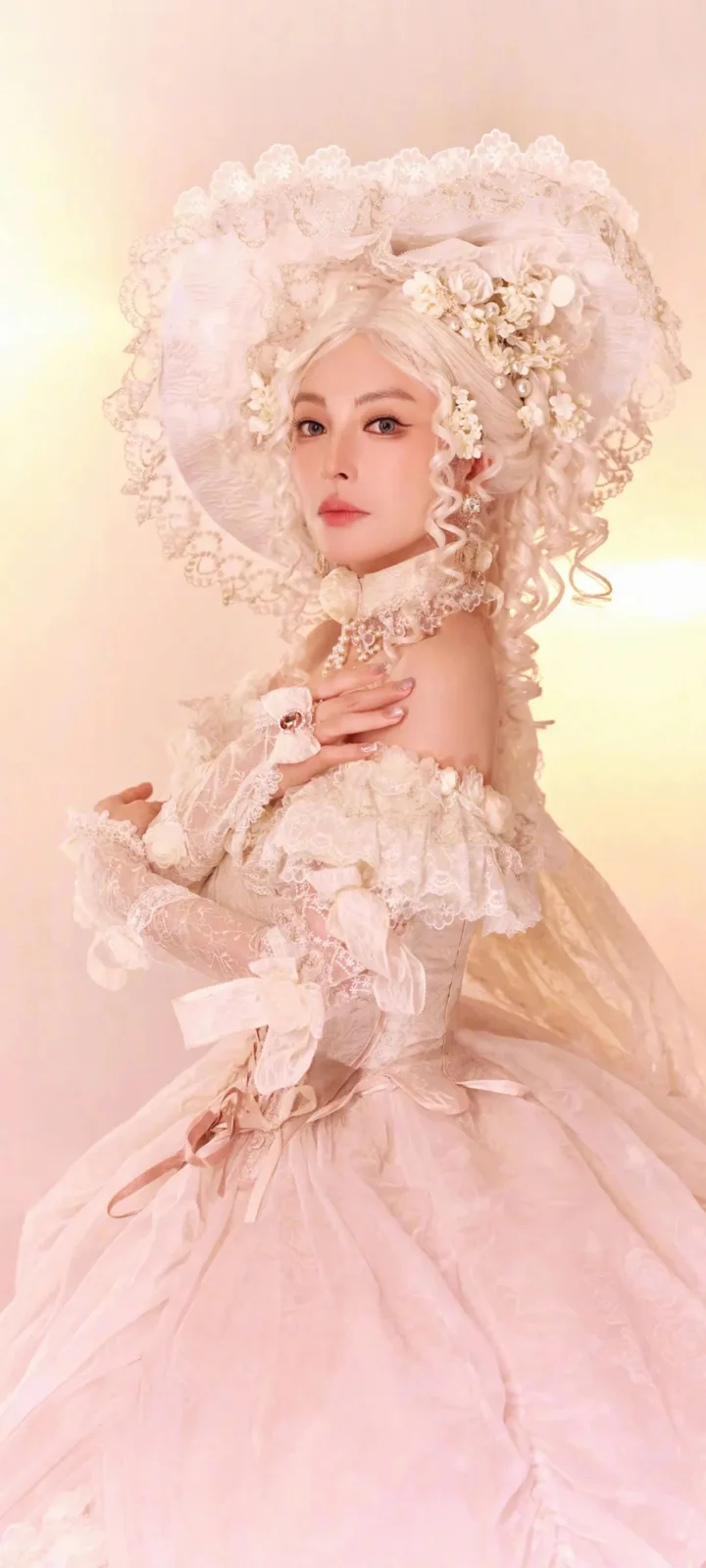 淡いピンクの華やかなドレスと花飾りのヘッドピースを身につけた女性が、柔らかなピンク色の背景の前でポーズを取っています。