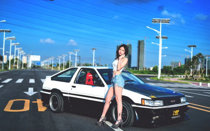 一名成年女性站在白色与黑色汽车旁，背景是晴朗蓝天、宽阔道路和成排街灯。
