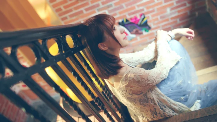 一名女性侧身坐在黑色长椅上，身后是砖墙。